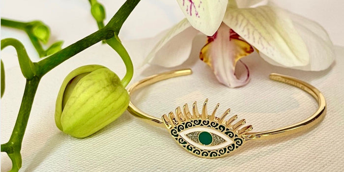 An evil eye bracelet with emerald green, white, and crystal enamel detailing shown with a flower over a canvas background.